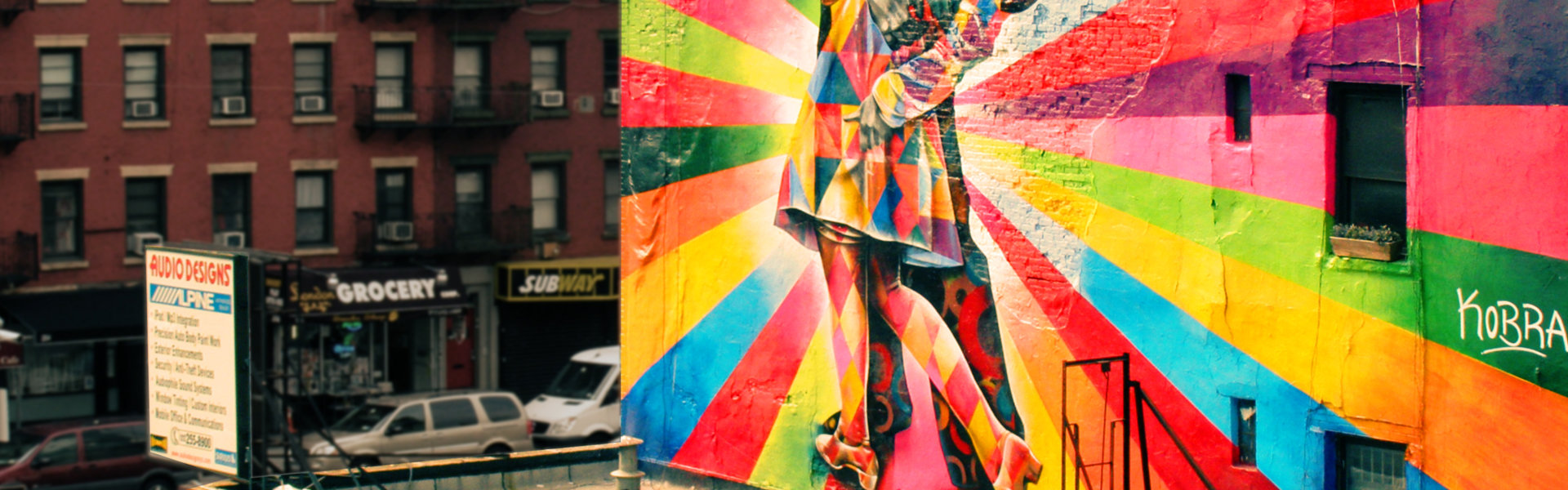 Colorful mural by artist Kobra depicting a couple kissing, set against a vibrant rainbow background in an urban area.